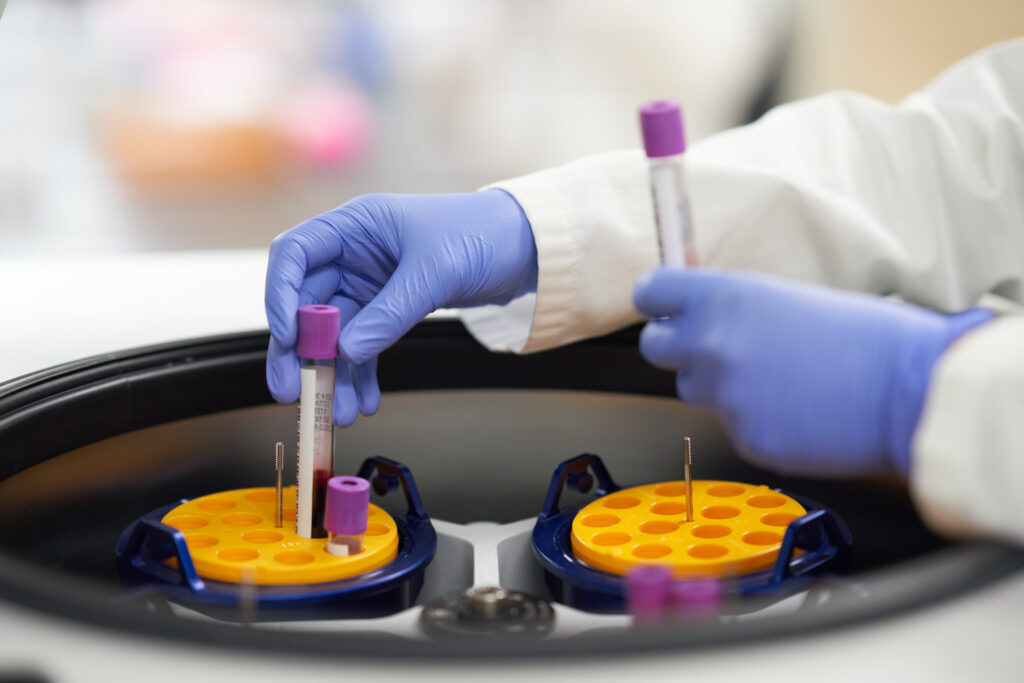 Gloved hands loading blood collection tubes into a centrifuge in the Biomarker Laboratory